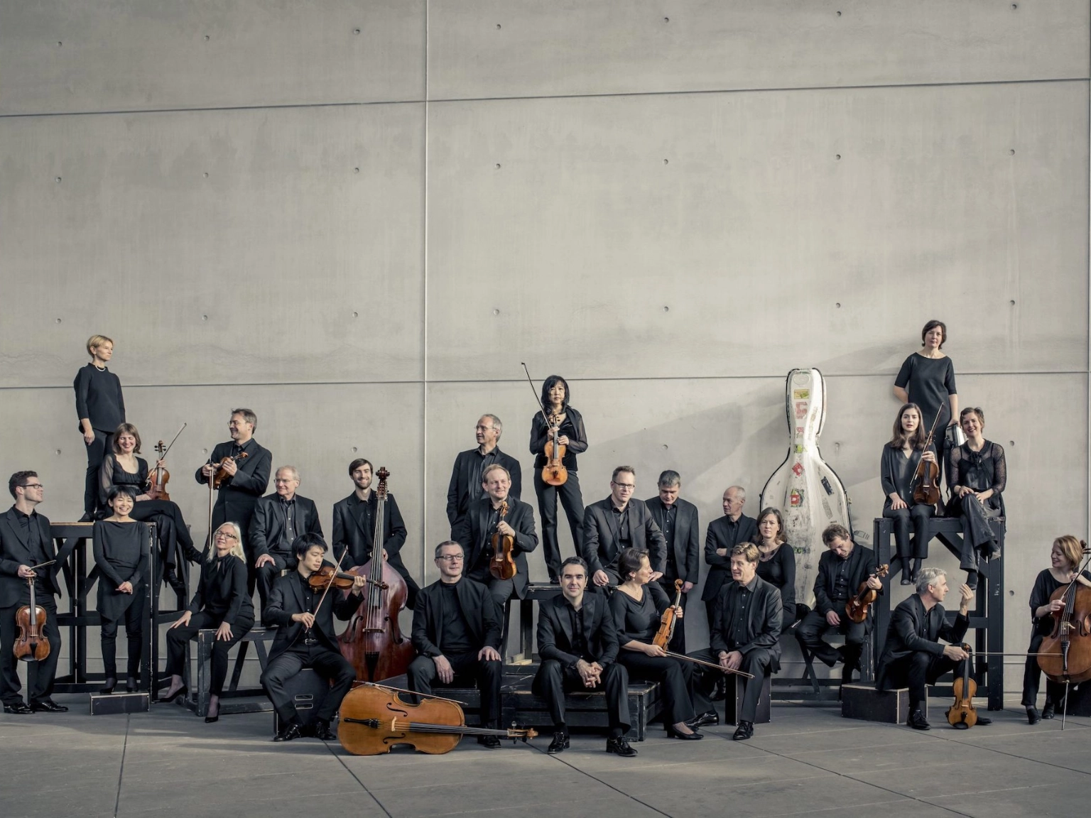 Das Münchener Kammerorchester vor einer grauen Betonwand. Sie posieren zusammen mit ihren Instrumenten. Manche schauen in die Kamera, andere unterhalten sich.