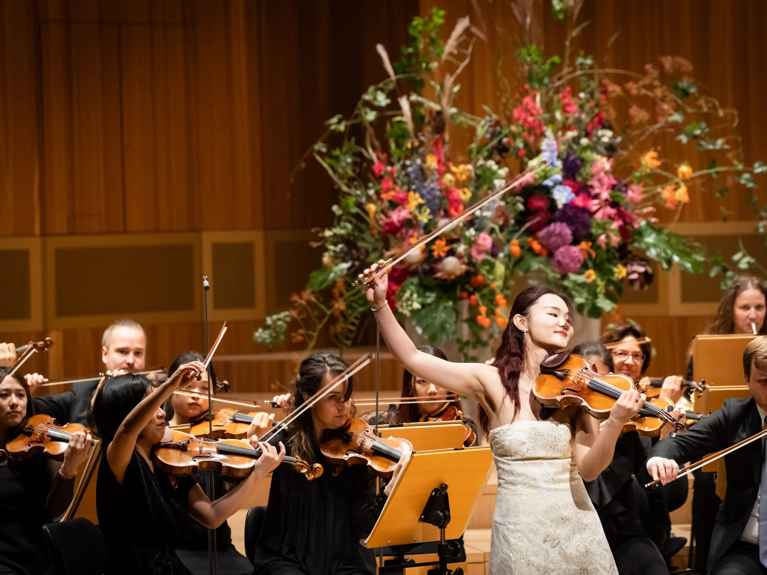Angela Chan steht in einem cremefarbenen Kleid auf der Bühne des Großen Saals im NDR Konzerthaus. Hinter ihr sind die Musiker*innen der NDR Radiophilharmonie und eine schöne Blumendekoration zu sehen. Sie hat soeben das Stück beendet, lächelt und hält die Violine in der linken Hand, den Bogen in der rechten, weit oben von ihr gestreckt. 